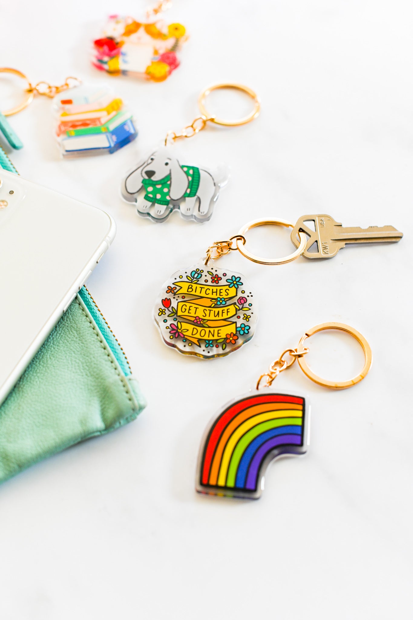 Collection of colorful acrylic keychains with gold rings including rainbow, Bitches Get Stuff Done, dog, book stack, and cat designs with styled mint green wallet on white background