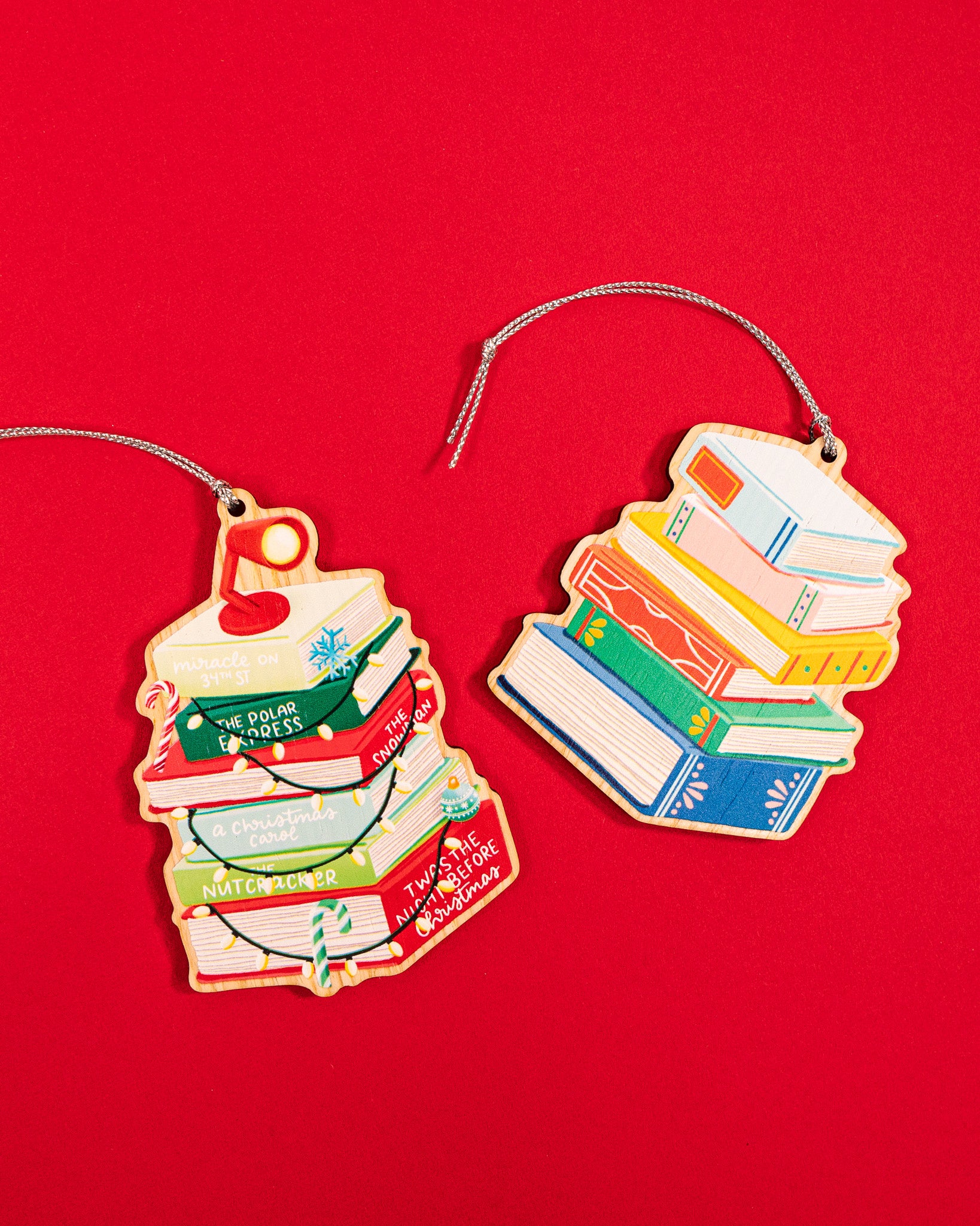 Two wood Christmas book ornaments with silver strings on red background - holiday classics with lights and colorful book stack 