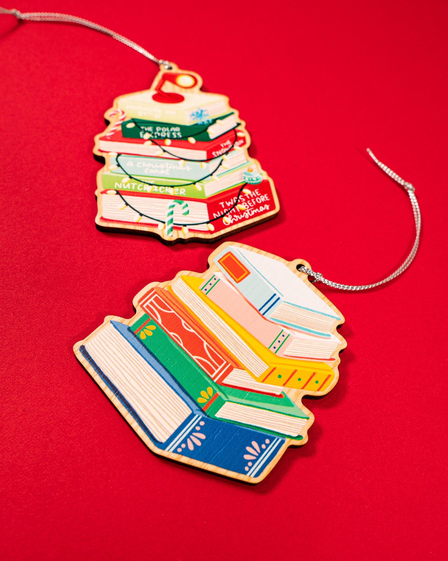 Close-up of two wooden book stack ornaments on vibrant red: Christmas classics featuring The Polar Express, The Snowman, Nutcracker, and Twas the Night Before Christmas with red desk lamp, candy cane, and string lights; plus colorful general book stack in blue, pink, yellow, orange, and green