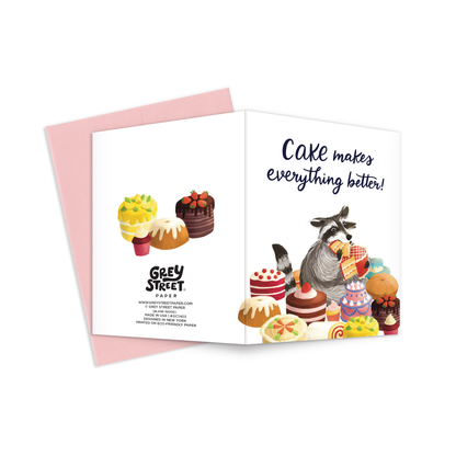 Full view of greeting card with illustration of a cute raccoon eating cake on the font and assorted cakes on the back with a pink envelope.