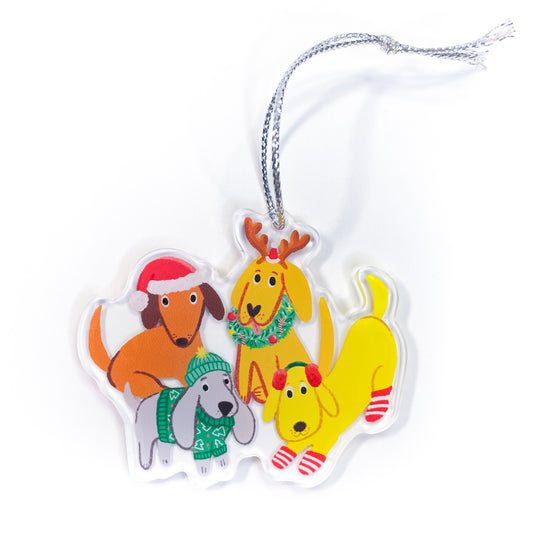 Holiday dogs acrylic ornament featuring four cute dogs in Christmas outfits: orange dachshund wearing red Santa hat with white trim, golden yellow dog with brown reindeer antlers and green wreath with red berries, gray dog wearing green knit scarf with white snowflakes, and bright yellow dog wearing red and white striped socks with silver hanging string 