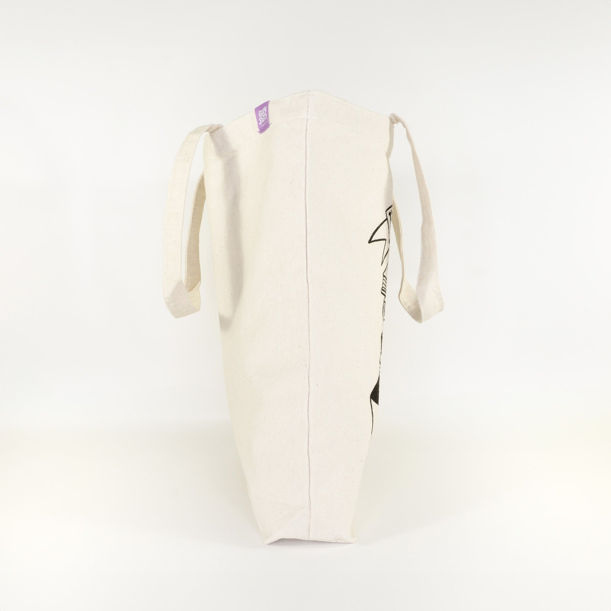 Side view of I'd Rather Be Reading cotton tote bag showing partial book stack design and Grey Street Paper purple label tag on natural canvas