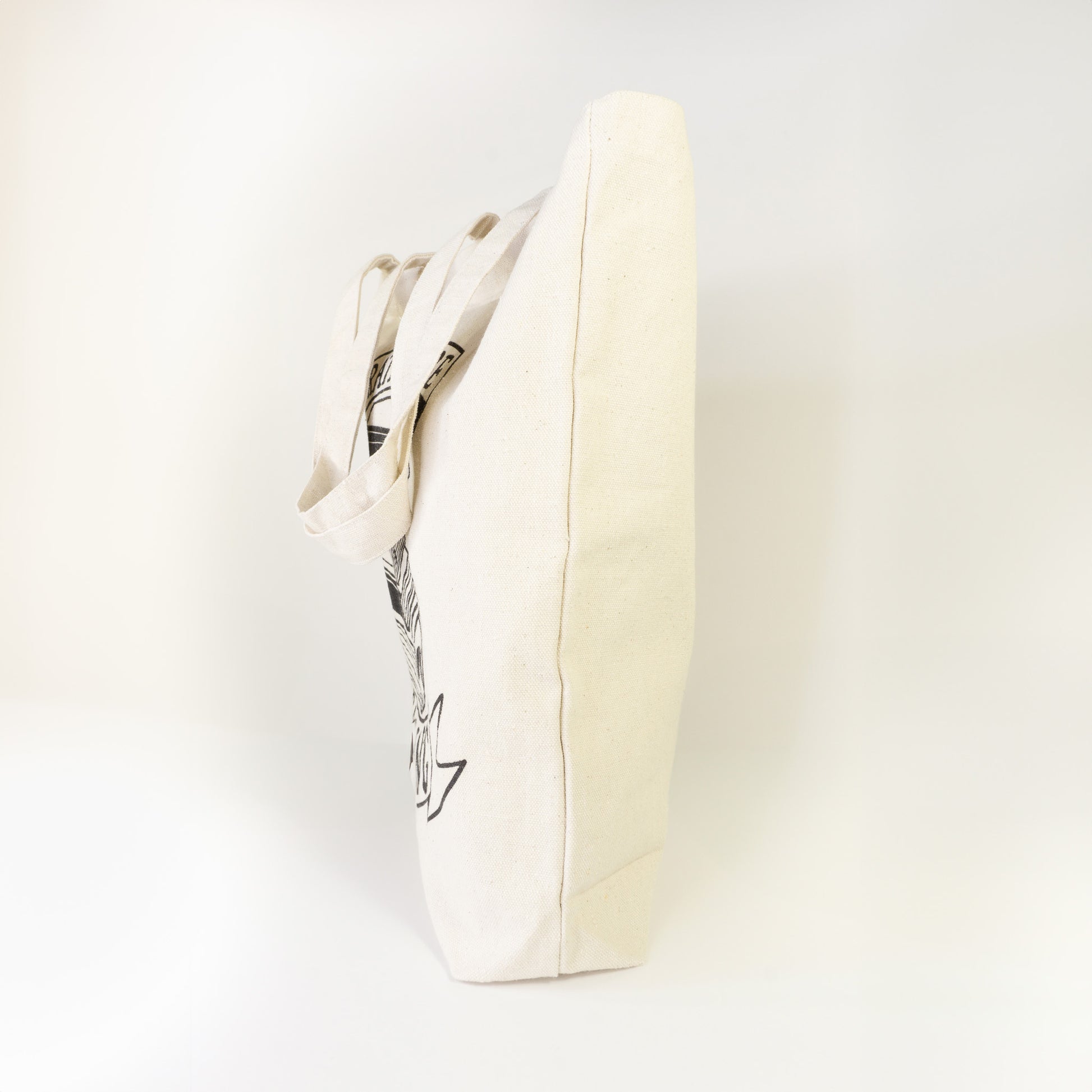 Side profile of I'd Rather Be Reading cotton tote bag showing edge of book illustration and ribbon banner on natural canvas