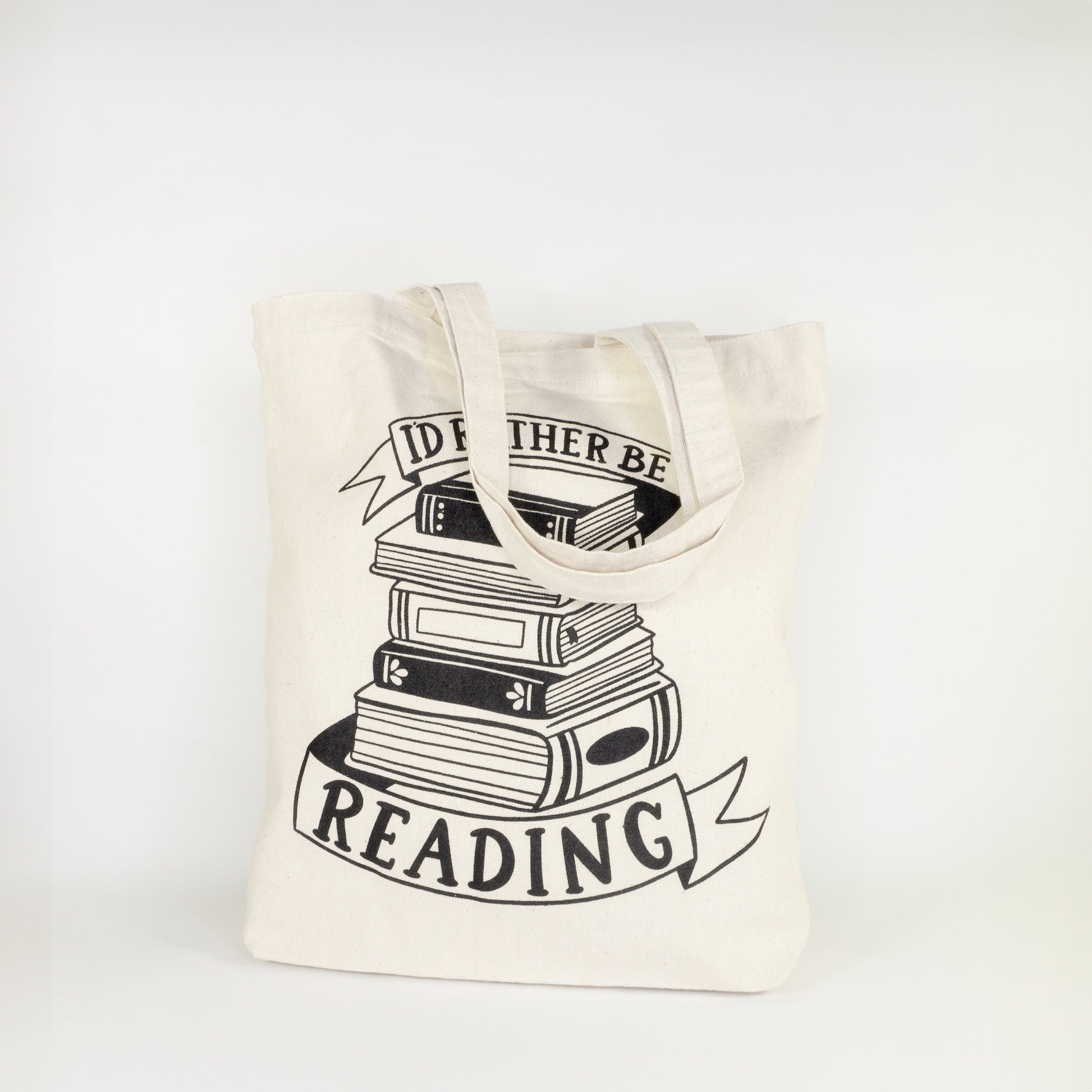 Angled view of I'd Rather Be Reading cotton tote bag with complete stack of books and ribbon banners design on natural canvas with shoulder straps