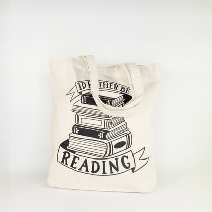 Angled view of I'd Rather Be Reading cotton tote bag with complete stack of books and ribbon banners design on natural canvas with shoulder straps
