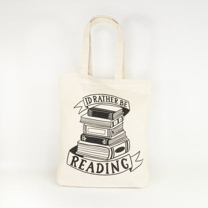 Front view of I'd Rather Be Reading cotton tote bag laying flat showing complete design with stack of books between ribbon banners on natural canvas