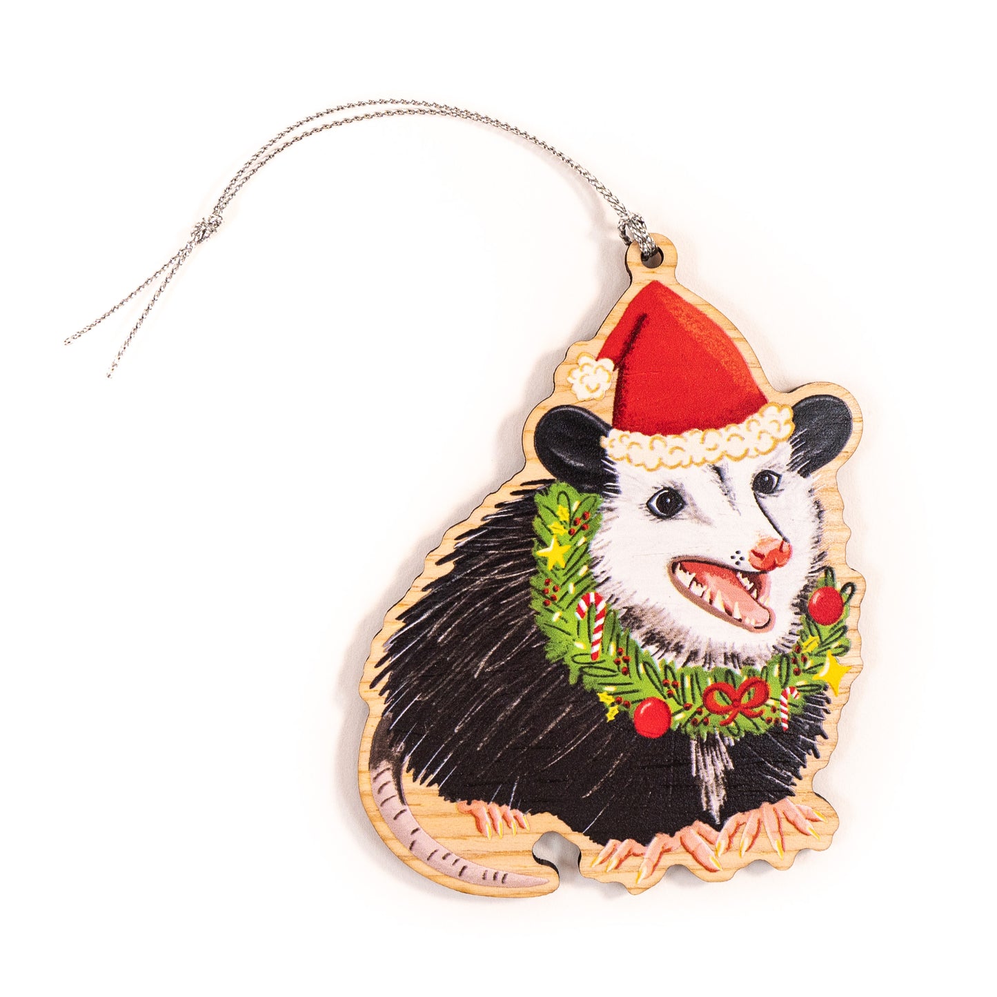Wooden Christmas ornament featuring smiling black and white opossum with pink open mouth wearing red Santa hat with white fur trim and pom pom, decorated with green evergreen wreath with red berries, red and white candy cane, and yellow flowers on natural birch wood with silver hanging string
