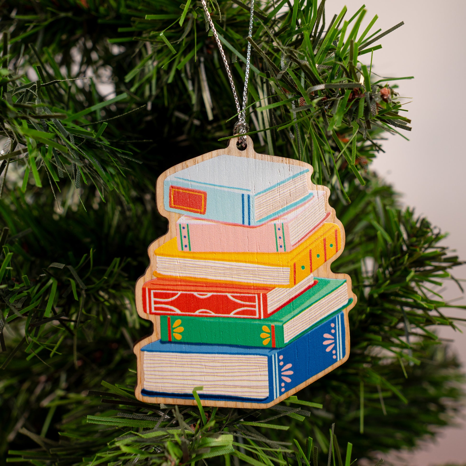 Colorful stack of books wood Christmas ornament with silver string hanging on evergreen tree featuring blue, pink, yellow, orange, and green hardcover books 