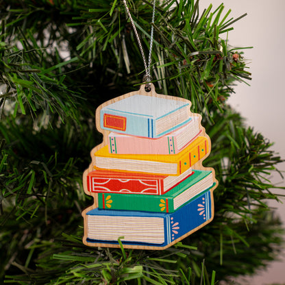 Colorful stack of books wood Christmas ornament with silver string hanging on evergreen tree featuring blue, pink, yellow, orange, and green hardcover books 