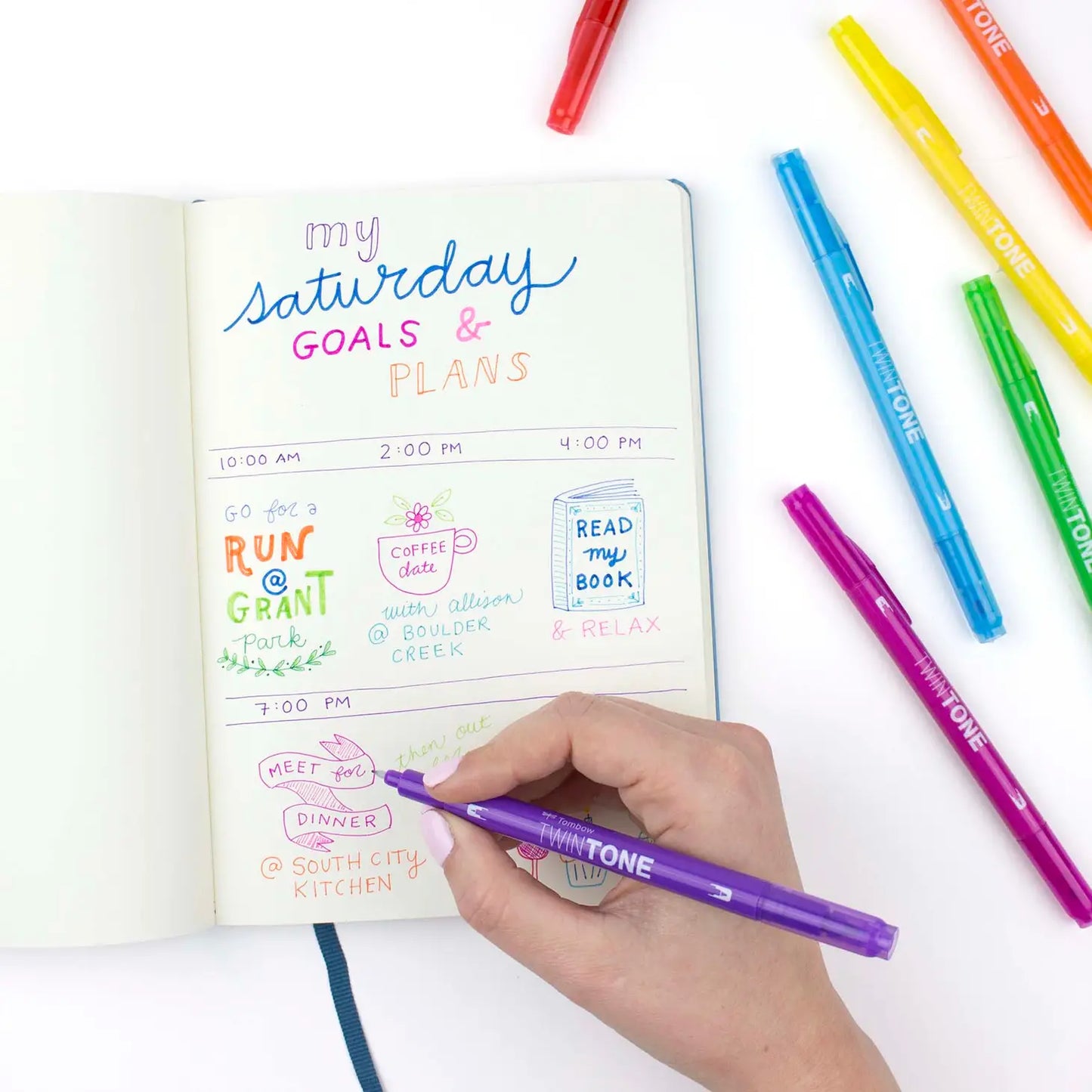 A stylized photo of a hand using a Tombow Twintone Marker to write Saturday Goals and Plans in a journal with Twin Tone Markers next to the journal.